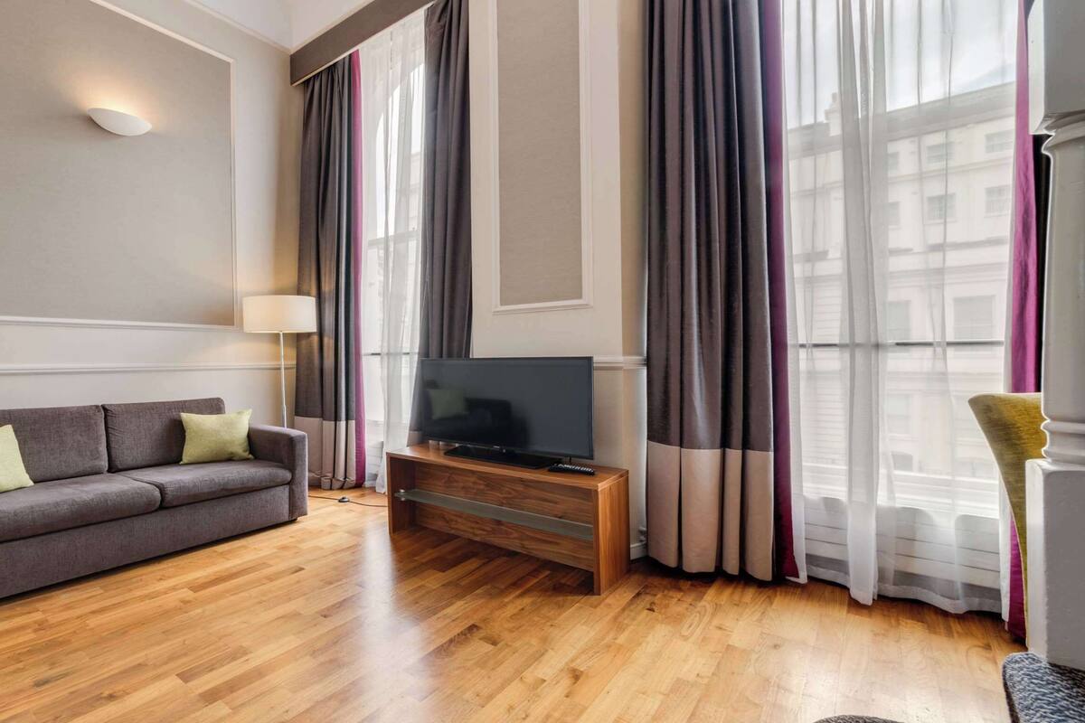 A modern living room with a grey sofa, wooden flooring, a TV on a wooden stand, and large windows with sheer curtains.