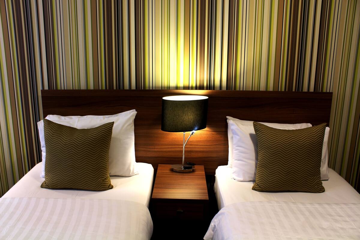 A hotel room with two beds, a nightstand with a lamp, and a striped wall.
