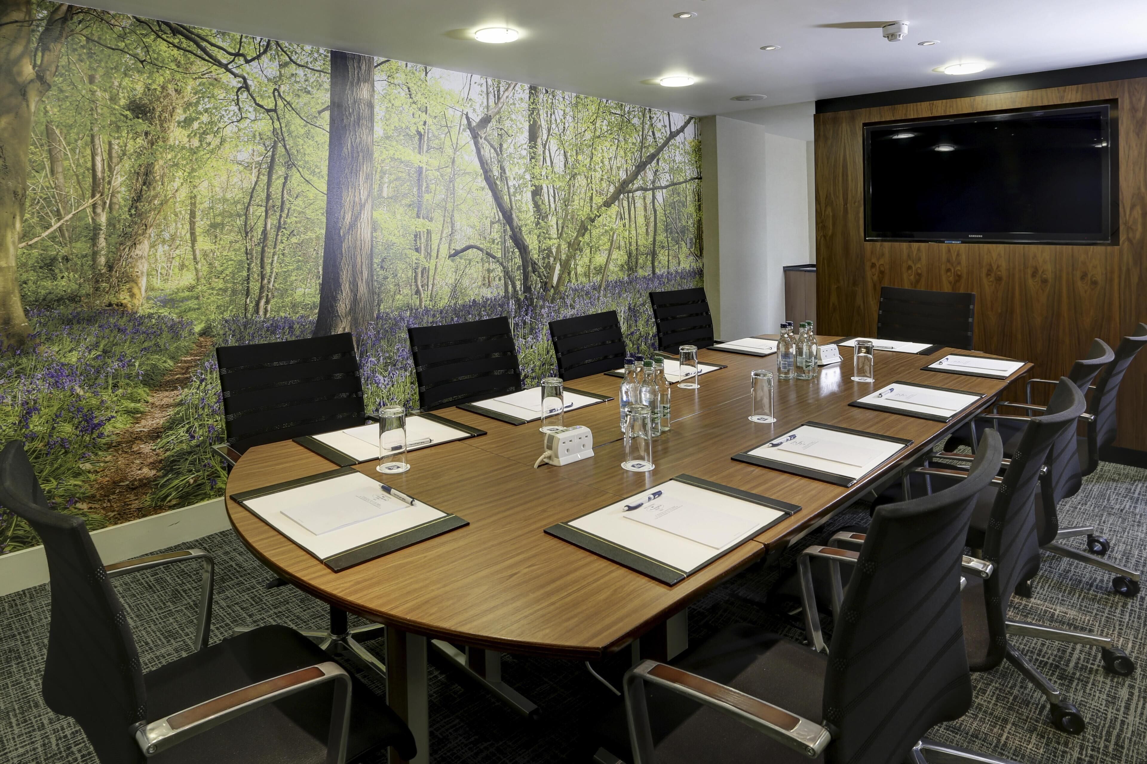 A conference room with a long wooden table, black chairs, and a large screen on the wall.