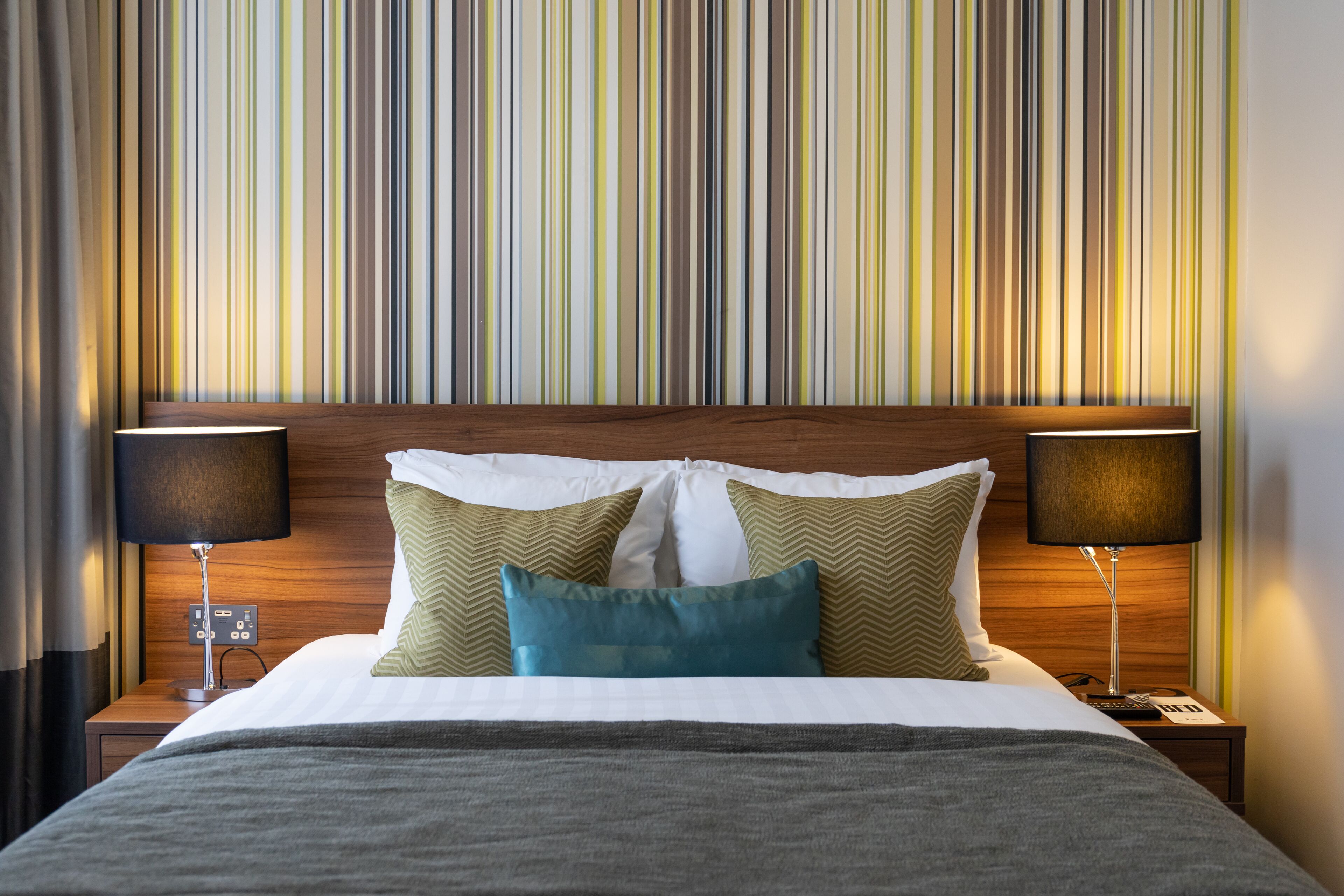 Una camera d'albergo con carta da parati a righe, una testiera in legno, un letto con cuscini bianchi e color acquamarina e due lampade da comodino.