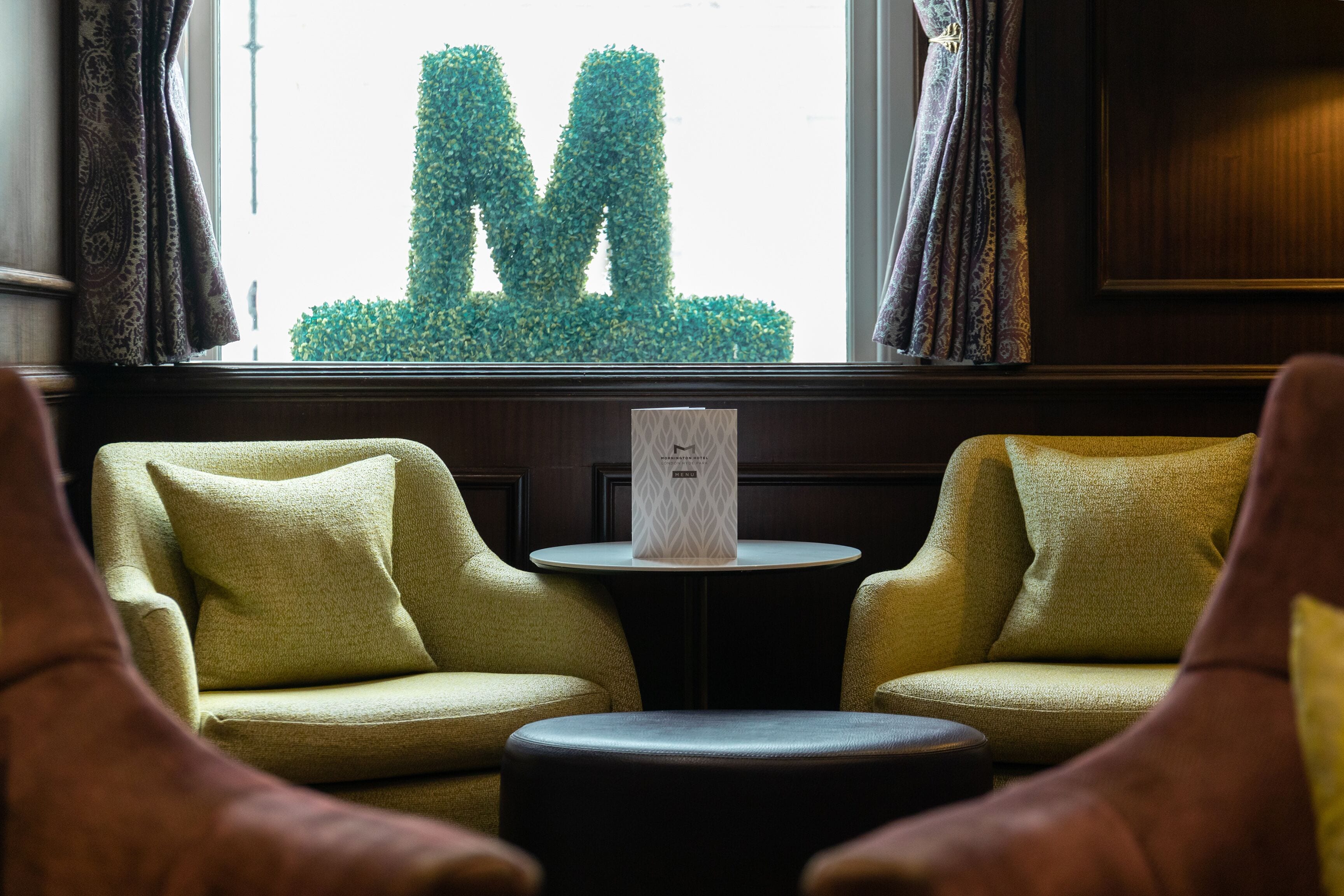 Una habitación con dos sillones amarillos, una pequeña mesa redonda con un objeto decorativo y vista a un seto que forma la letra 'M' a través de la ventana.