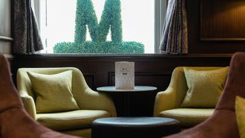 A room with two yellow armchairs, a small round table with a decorative item, and a view of a hedge forming the letter 'M' through the window.