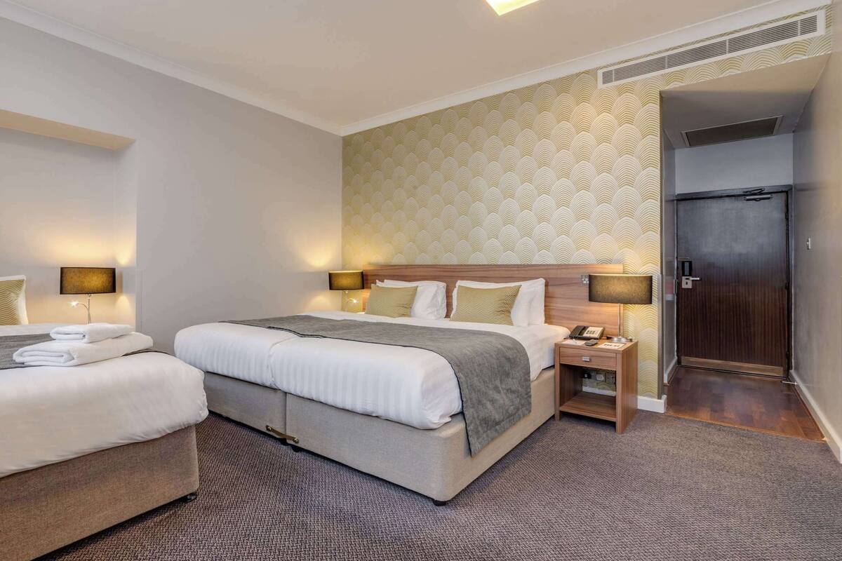 A hotel room with two beds, a wooden headboard, bedside tables with lamps, a small desk with a telephone, and a door leading to a bathroom.