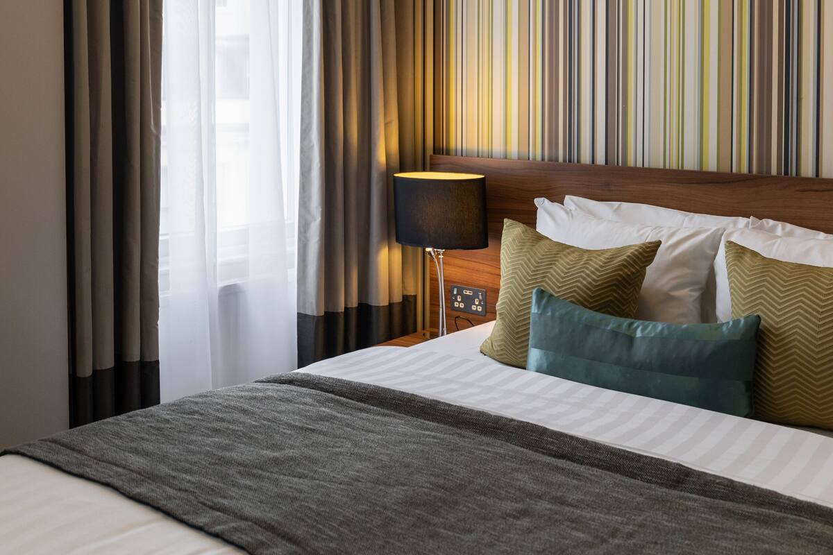 A hotel room with a large bed, a headboard, a bedside table with a lamp, and a striped wall.