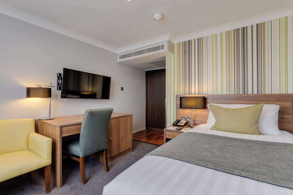 A hotel room with a bed, a desk with a chair, a television, and a bedside table with a lamp.