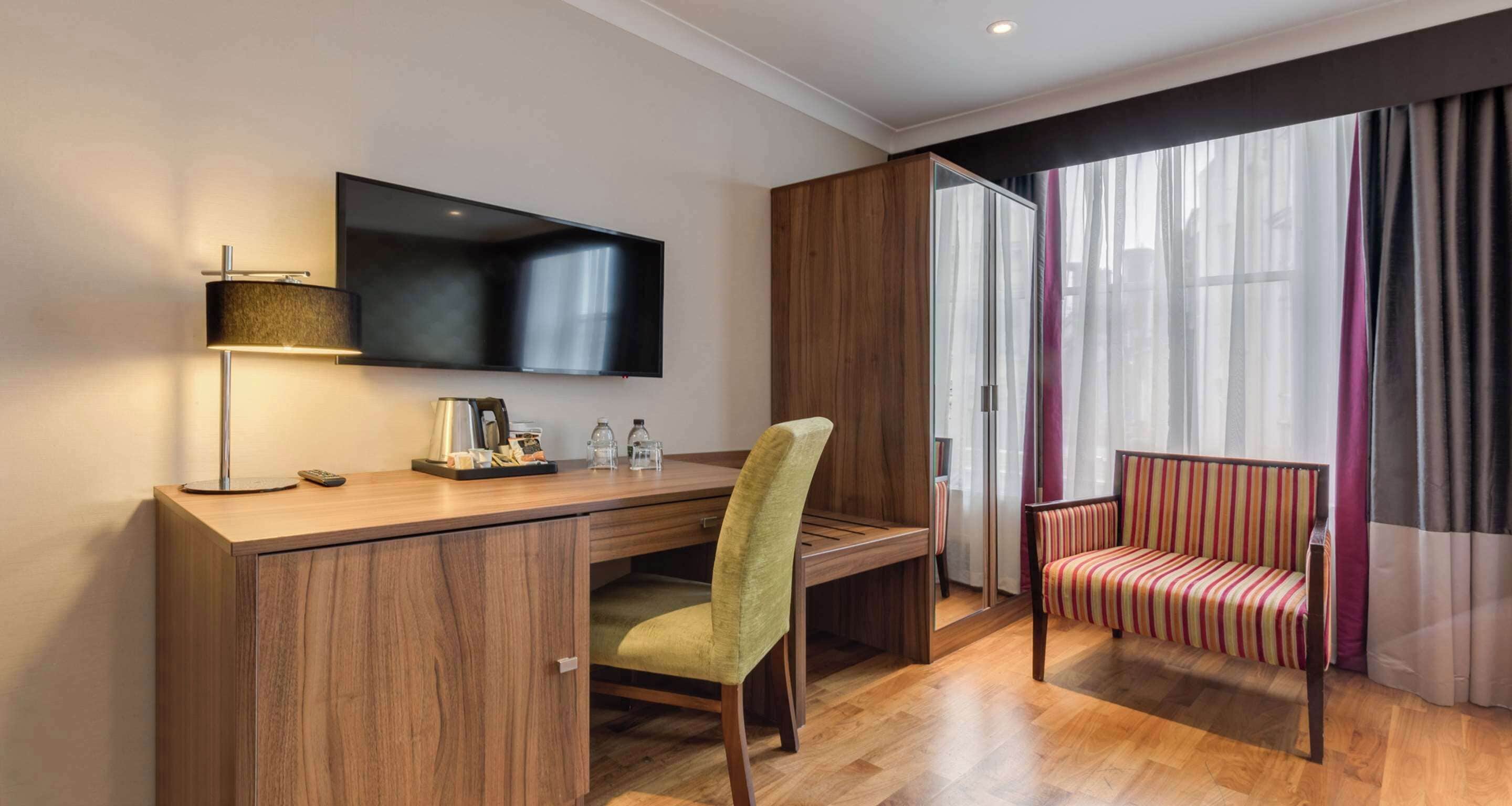 Hotelkamer met een houten bureau, een groene stoel, een aan de muur bevestigde televisie, een bank met gestreepte kussens en een groot raam met dunne gordijnen.