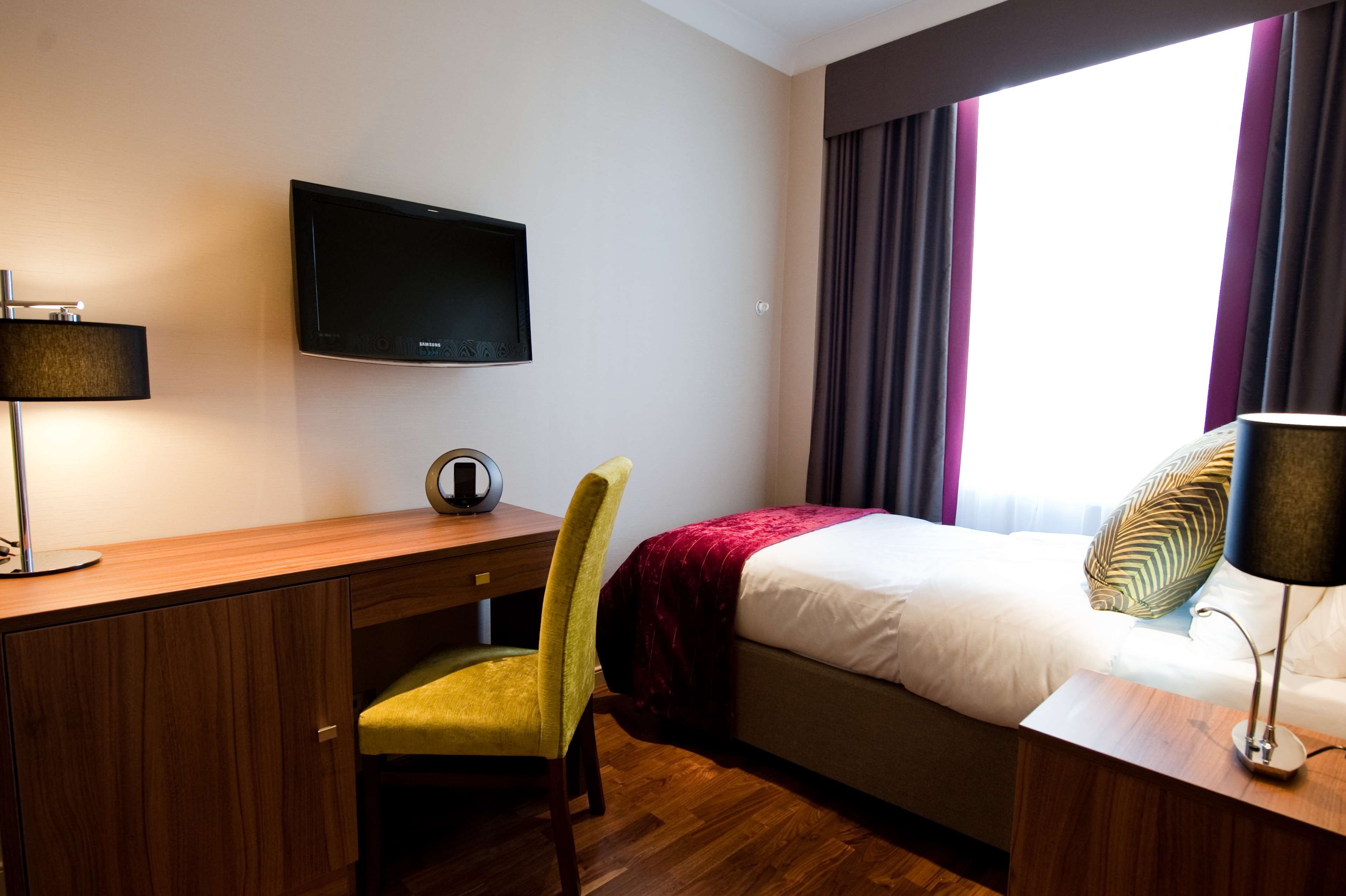 Habitación de hotel con cama, escritorio, silla, televisor y ventana con cortinas.