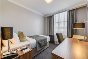 Ein Hotelzimmer mit Bett, Schreibtisch, Stuhl, Lampe und einem fenster mit Vorhängen.