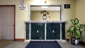 Un área de recepción con un mostrador de mármol, un felpudo azul, un certificado enmarcado, una planta y un cartel que indica internet de alta velocidad gratuito y habitaciones para fumadores.