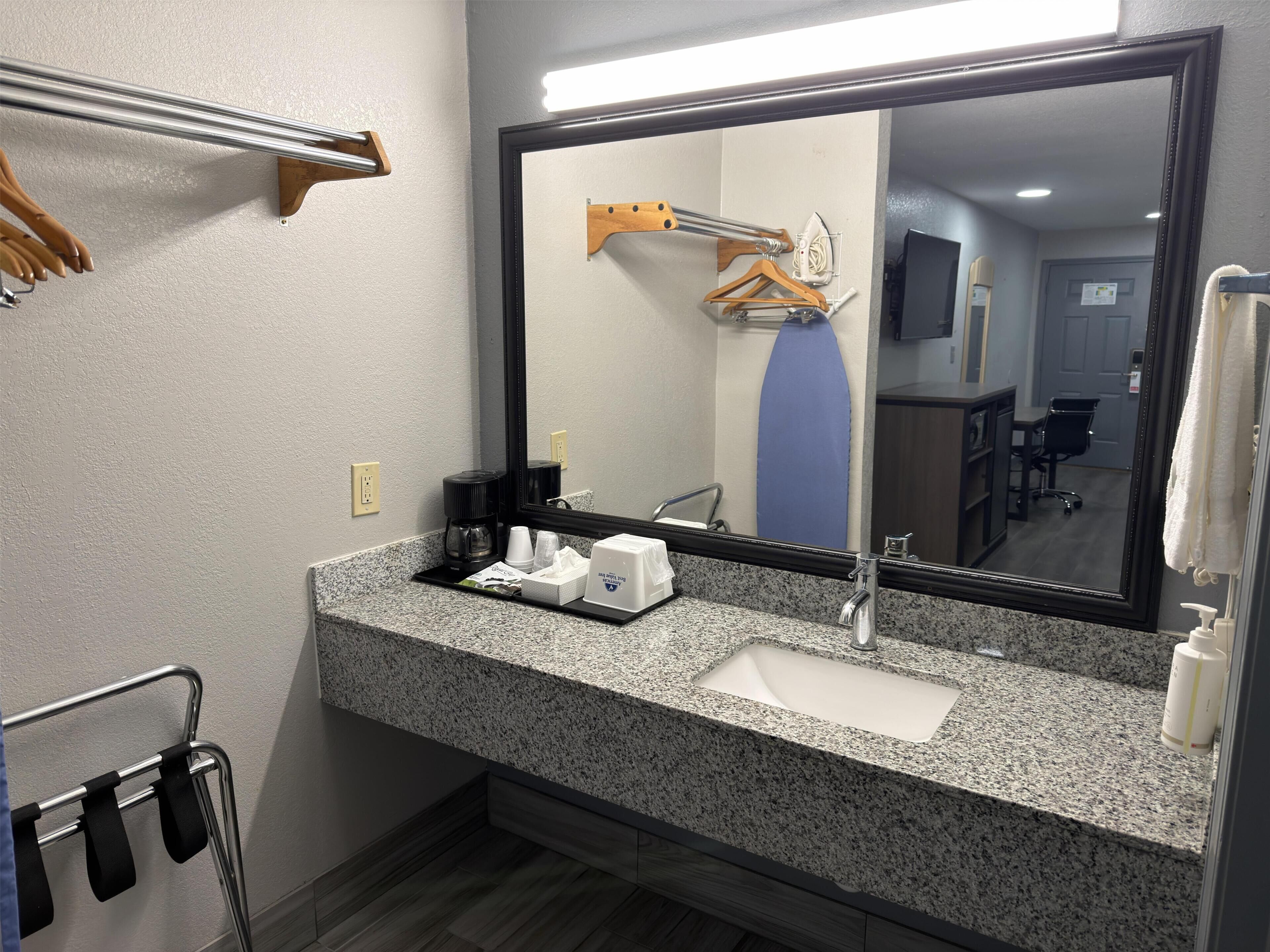 A hotel bathroom with a granite countertop, a sink, a mirror, a towel rack, a shower curtain, and a coffee maker.