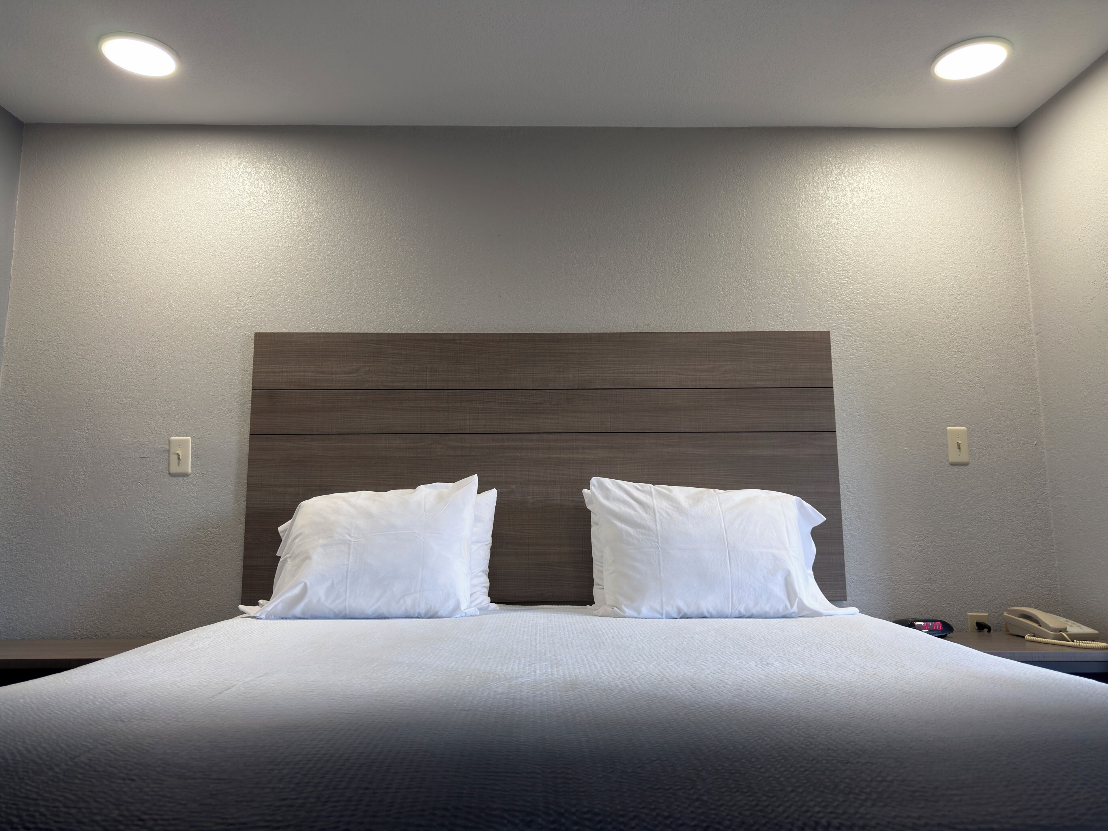 Hotelkamer met een bed, twee witte kussens, een houten hoofdbord, een nachtkastje met een telefoon en een klok, en inbouwspots.