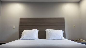 Hotelkamer met een bed, twee witte kussens, een houten hoofdbord, een nachtkastje met een telefoon en een klok, en inbouwspots.