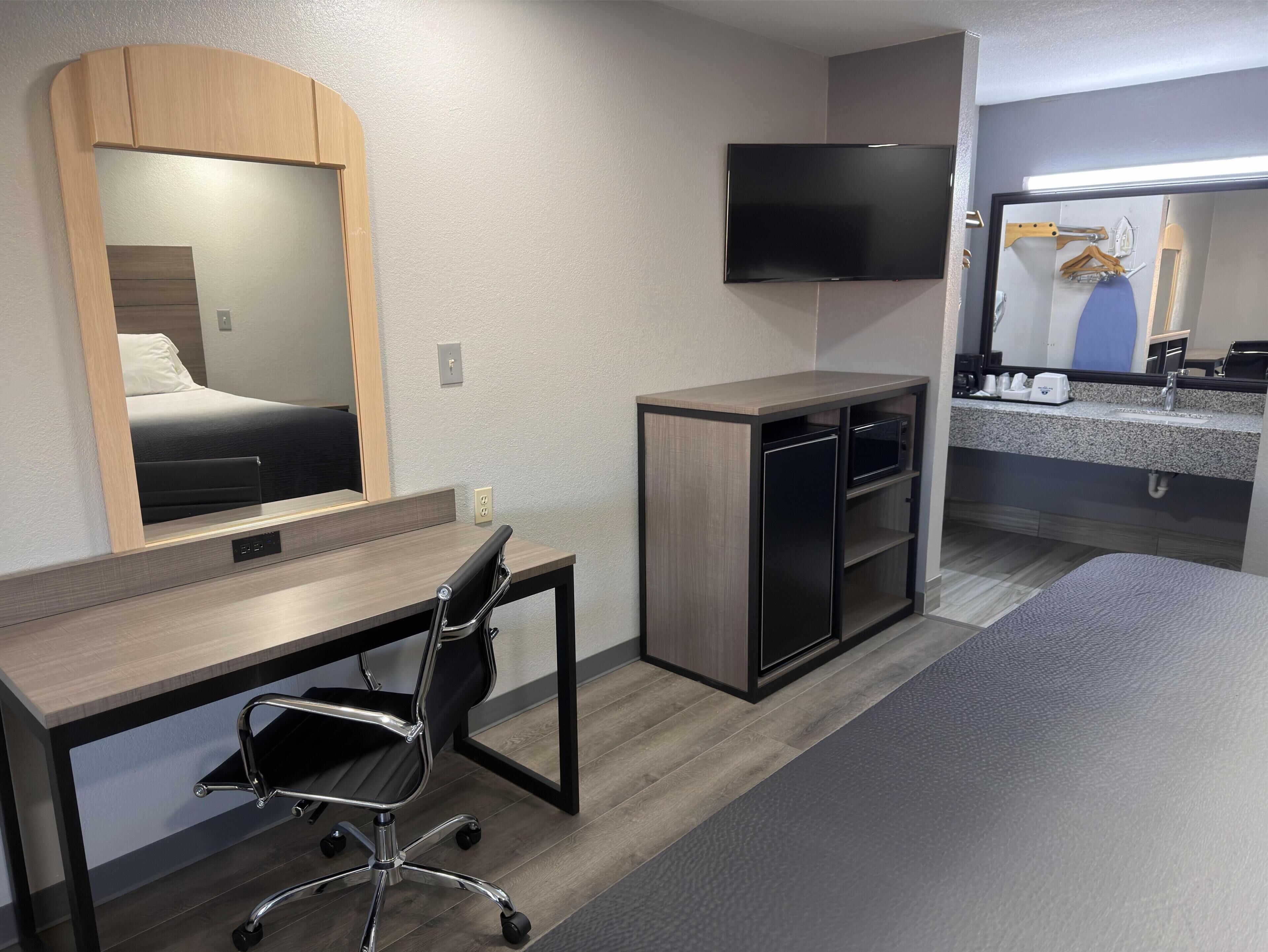 A hotel room with a bed, a desk with a chair, a television, a microwave, and a small kitchenette.