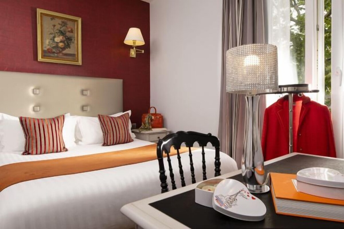 A hotel room with a bed, a dining table with a lamp, a chair, and a red coat hanging on the wall.