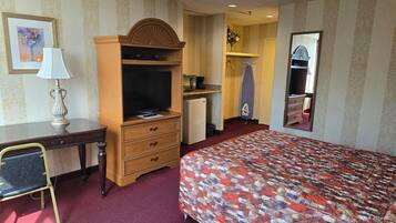 A hotel room with a bed, a desk with a lamp, a television, a small refrigerator, and a mirror.