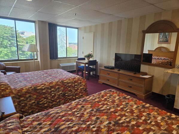 A hotel room with two beds, a desk, a TV, and a window with a view of buildings and trees. - Amsterdam Hotel (Stamford)