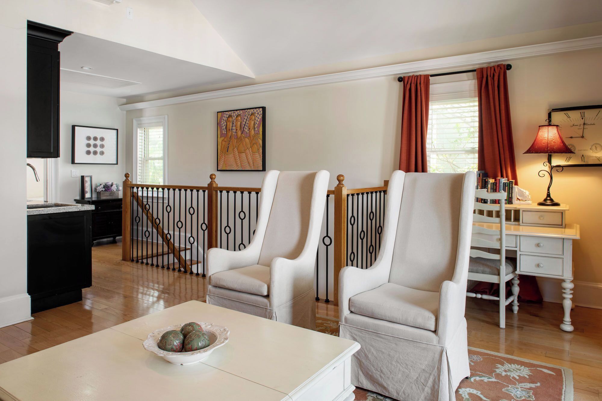 A spacious living room with two white armchairs, a coffee table with decorative items, a bookshelf, and a clock on the wall.