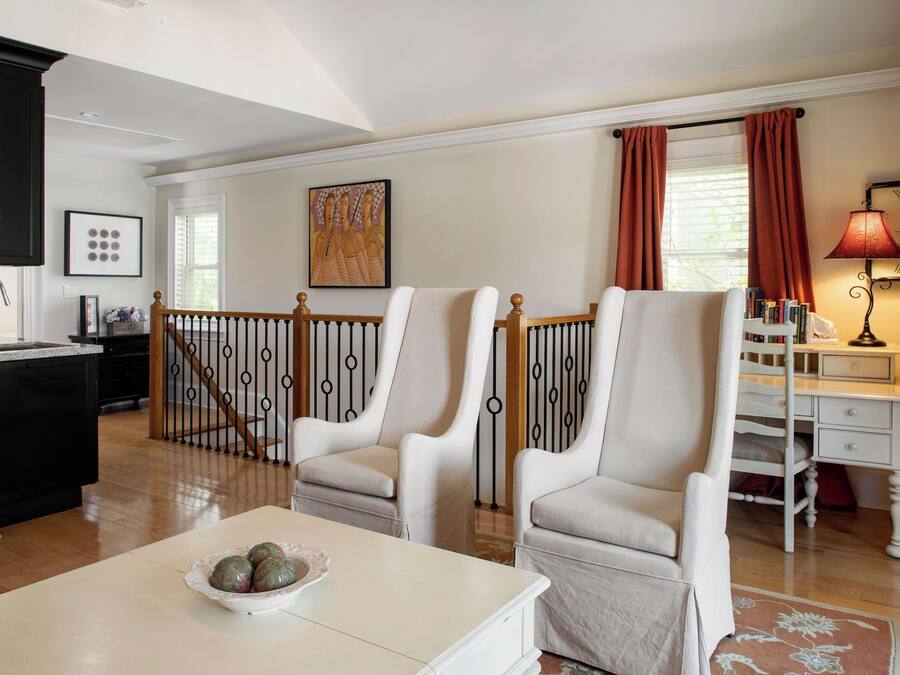 A spacious living room with two white armchairs, a coffee table with decorative items, a bookshelf, and a clock on the wall.