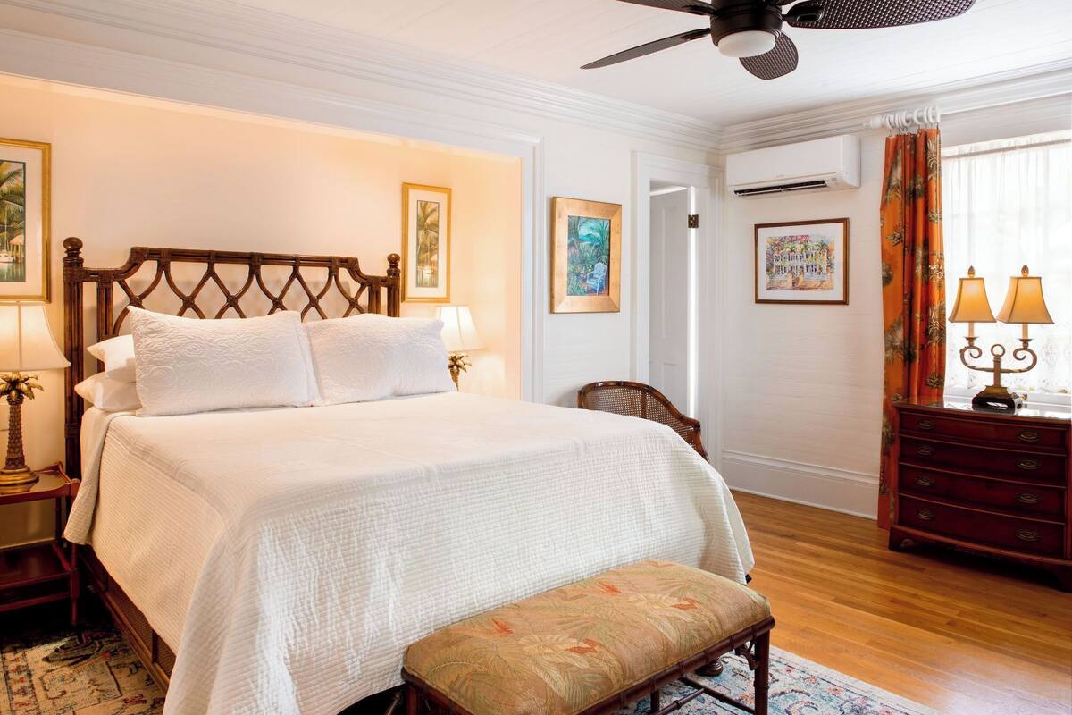 A neatly arranged bedroom with a large bed, a wooden bench, a patterned rug, and a ceiling fan.