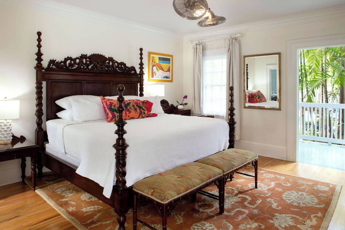 A four-poster bed with a bench, a wooden floor, a patterned rug, a painting, and a window with curtains.