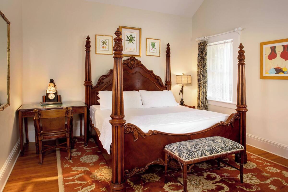A four-poster bed with a wooden headboard, a bedside table with a lamp, a bench, and a desk with a clock.