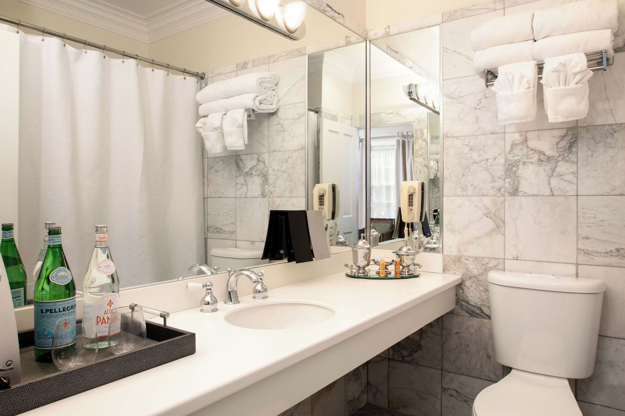 Une salle de bain moderne avec un pare-feu en marbre, un grand miroir, un lavabo blanc et des toilettes. Des serviettes sont pliées et empilées sur le mur, et une étagère en verre contient des bouteilles.
