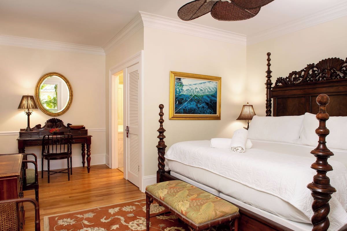 A bedroom with a four-poster bed, a desk with a chair, a bench, and a large rug.