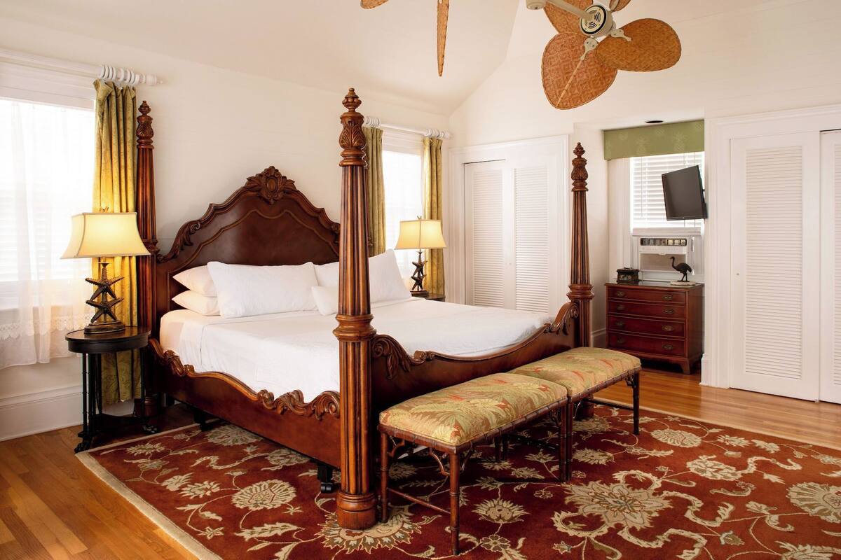 A four-poster bed with a wooden headboard, two bedside tables with lamps, a bench, and a wooden dresser.