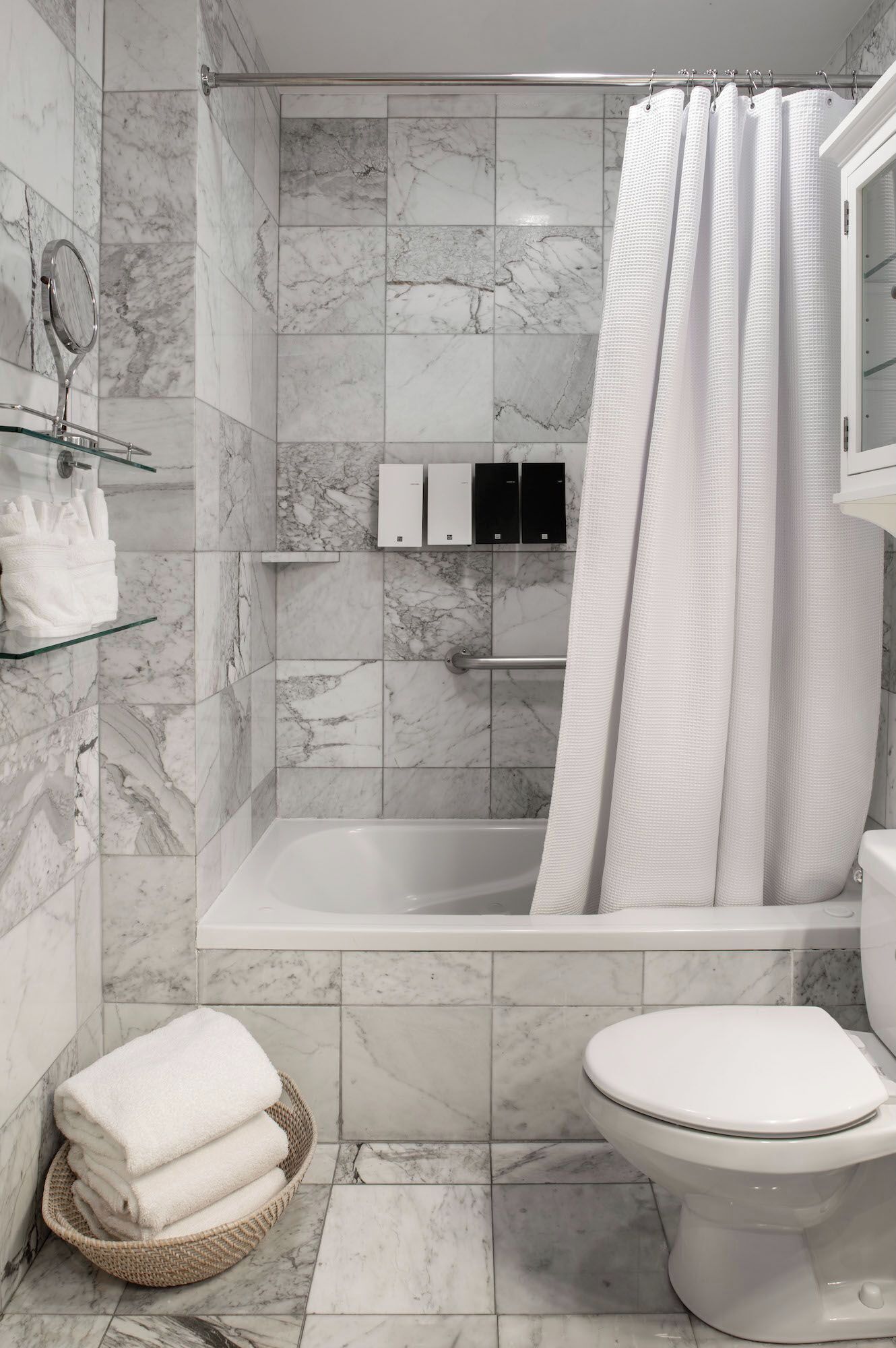 A modern bathroom with a marble-tiled shower, a glass shelf with toiletries, a toilet, and a basket of towels.