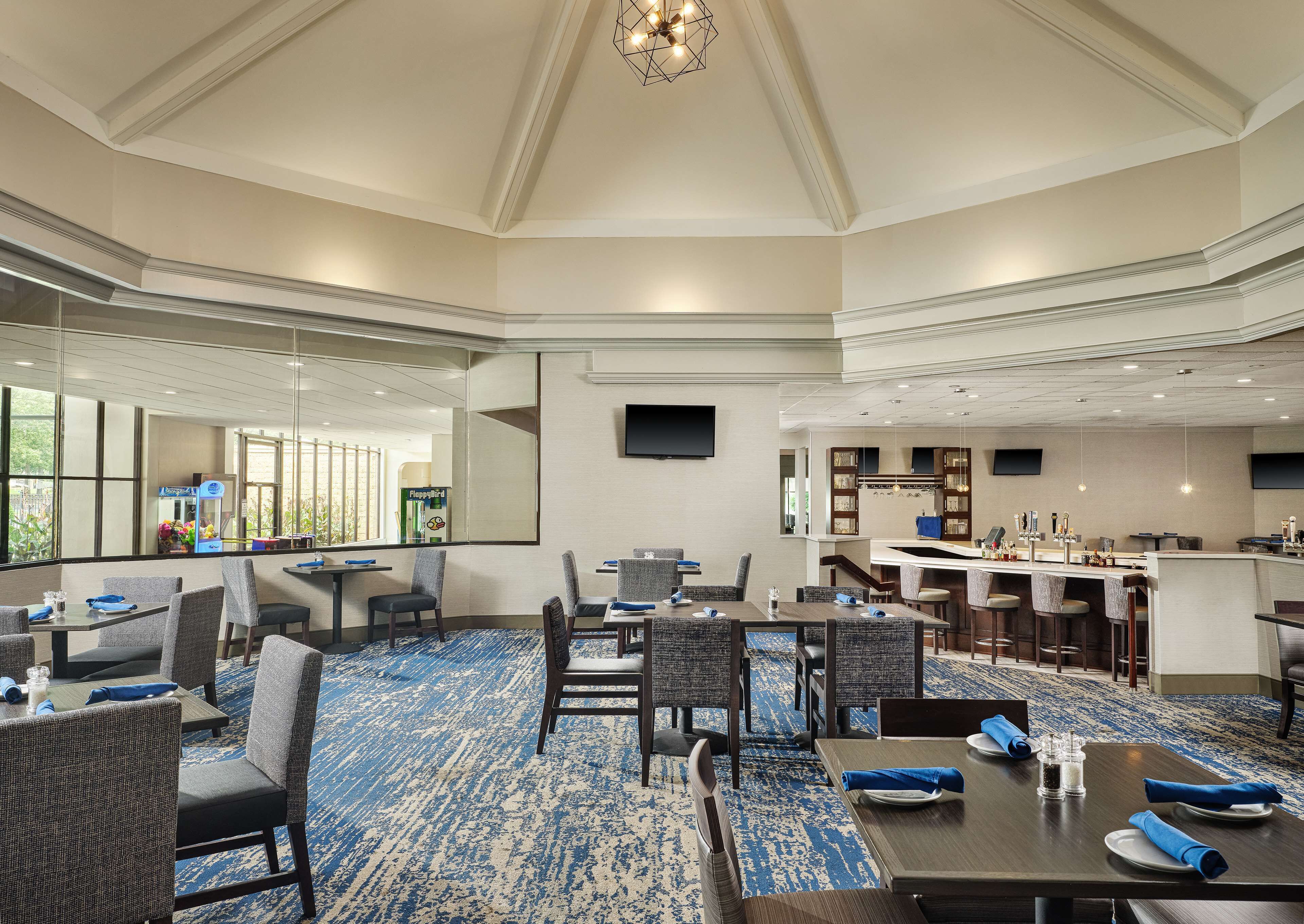 A hotel conference room with multiple tables and chairs, a bar area, and a large window providing a view of the outdoors.