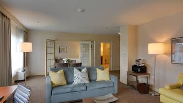 A hotel room with a grey sofa, yellow armchair, and a small table with a lamp. There is a kitchenette area with a microwave and a coffee maker.