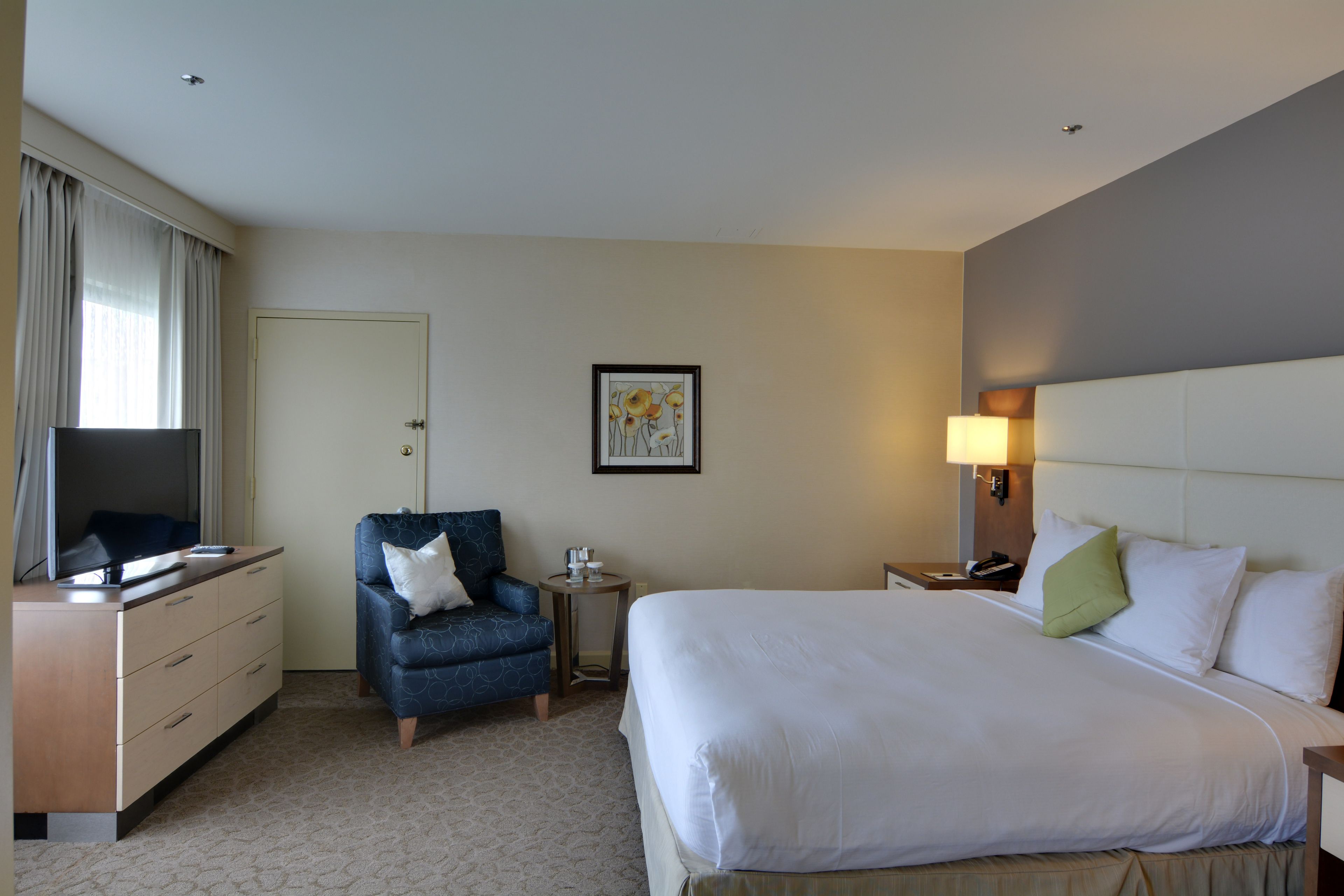 Habitación de hotel con cama, escritorio con televisor, una silla, mesita de noche y lámpara de pared.