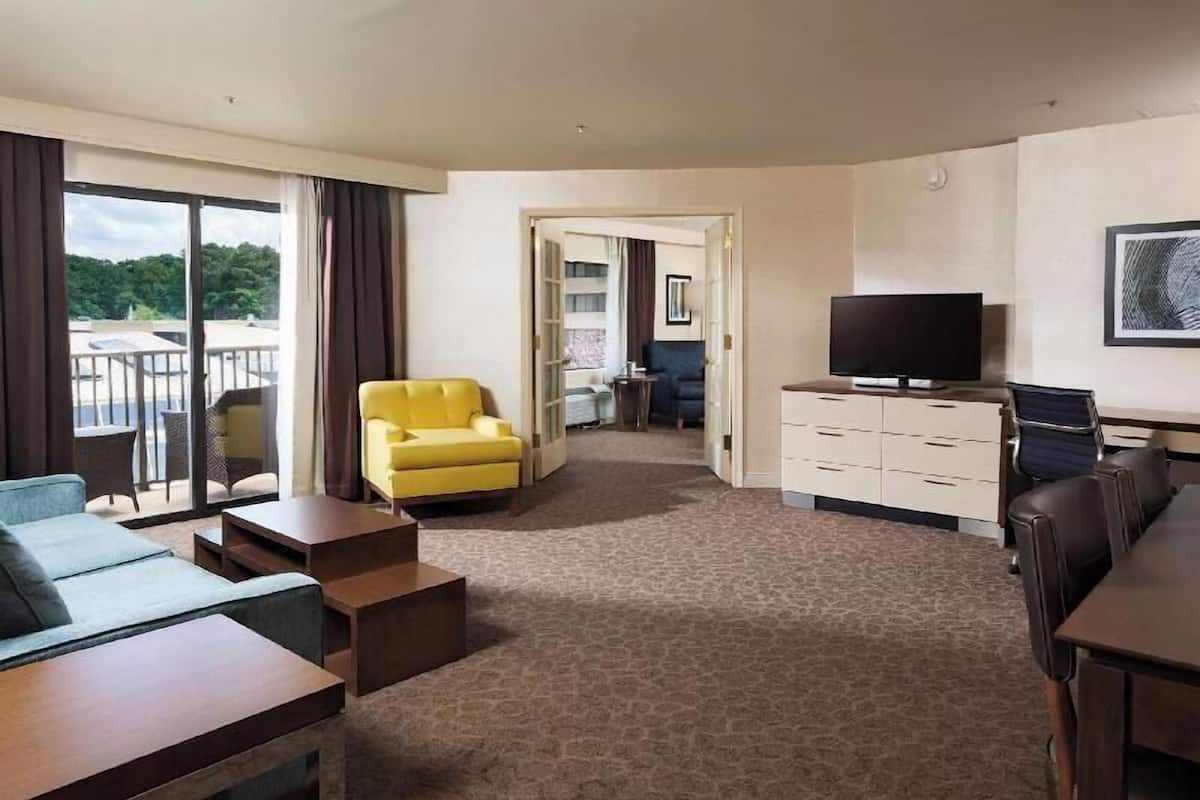 A hotel room with a sofa, a yellow armchair, a TV, a desk, and a view of the outdoors.