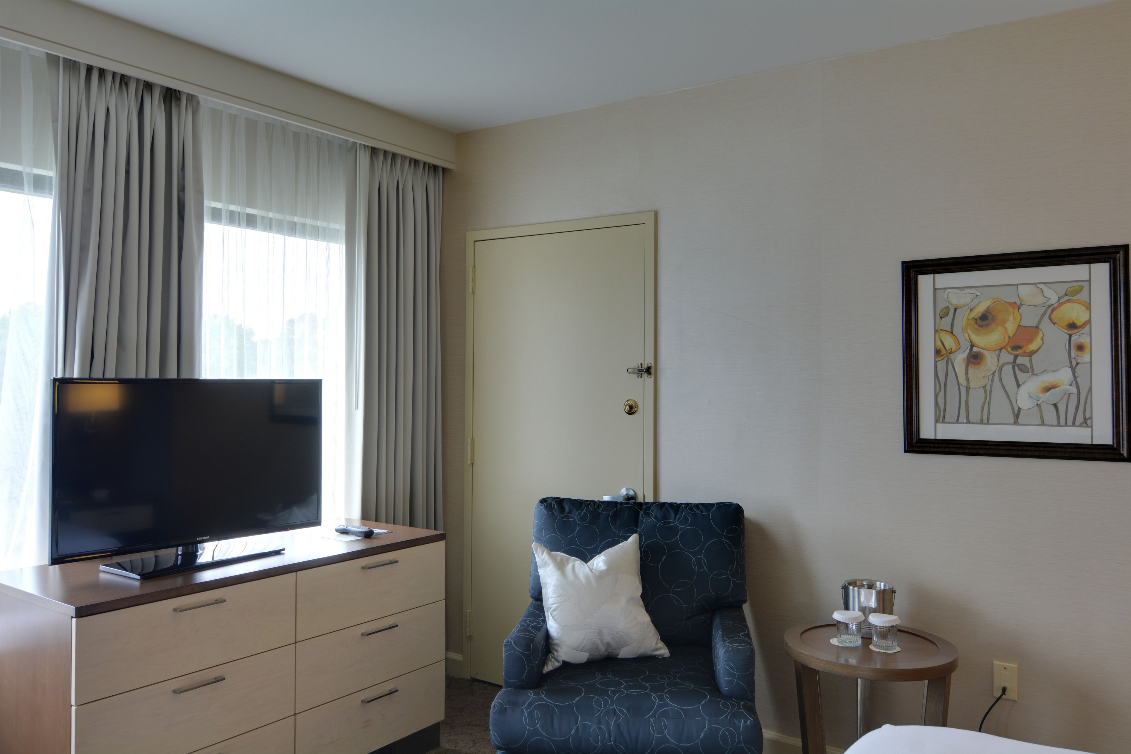 A hotel room with a flat-screen TV on a dresser, a blue armchair with a pillow, a small round table with a pitcher and glasses, and a framed floral artwork on the wall.