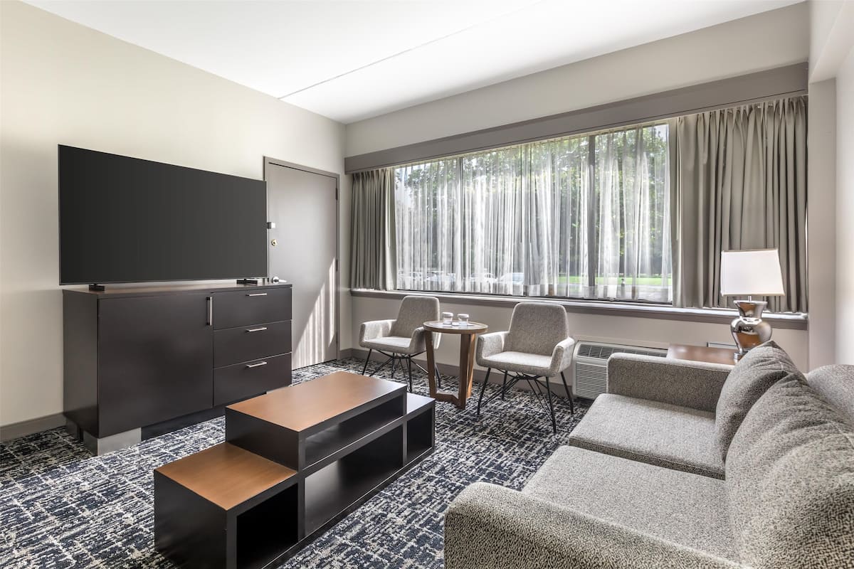 A hotel room with a flat-screen TV, a sofa, two armchairs, a small table, and a window with curtains.