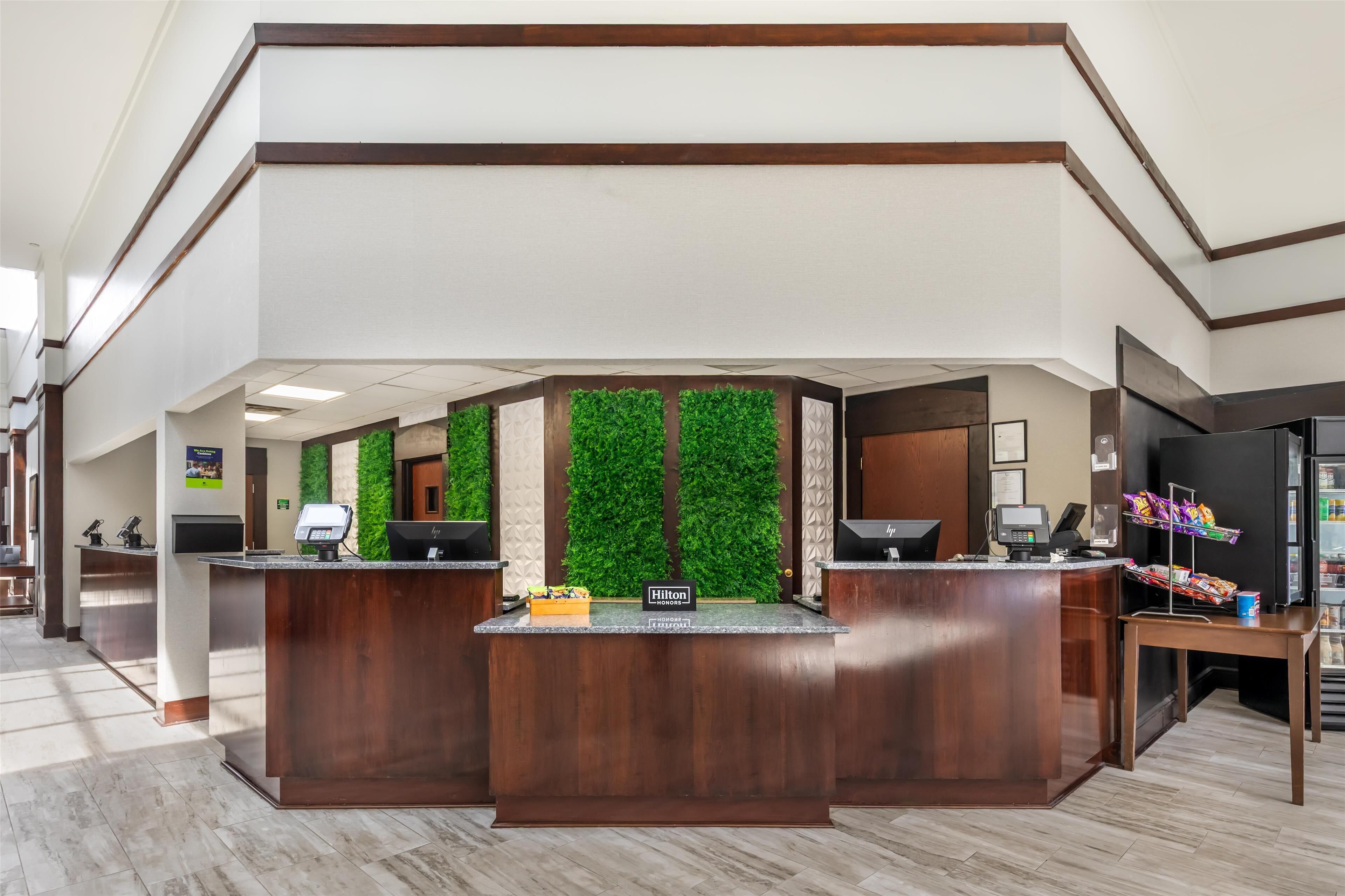 A hotel reception area with a wooden reception desk, a green vertical garden, and a self-service kiosk.