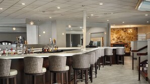 A modern bar area with a long counter, multiple stools, and a stone accent wall. - DoubleTree by Hilton Williamsburg (Williamsburg)