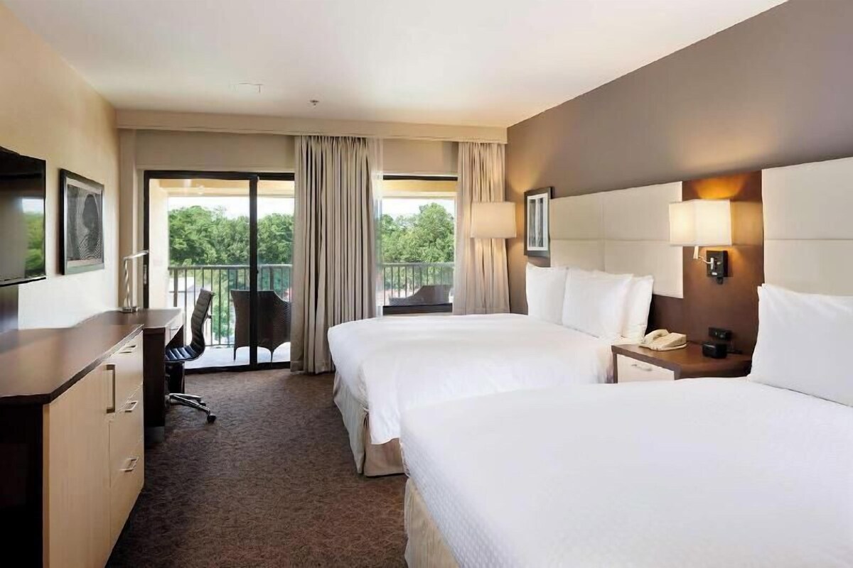 Habitación de hotel con dos camas, un escritorio, una silla, televisión y balcón con cortinas.