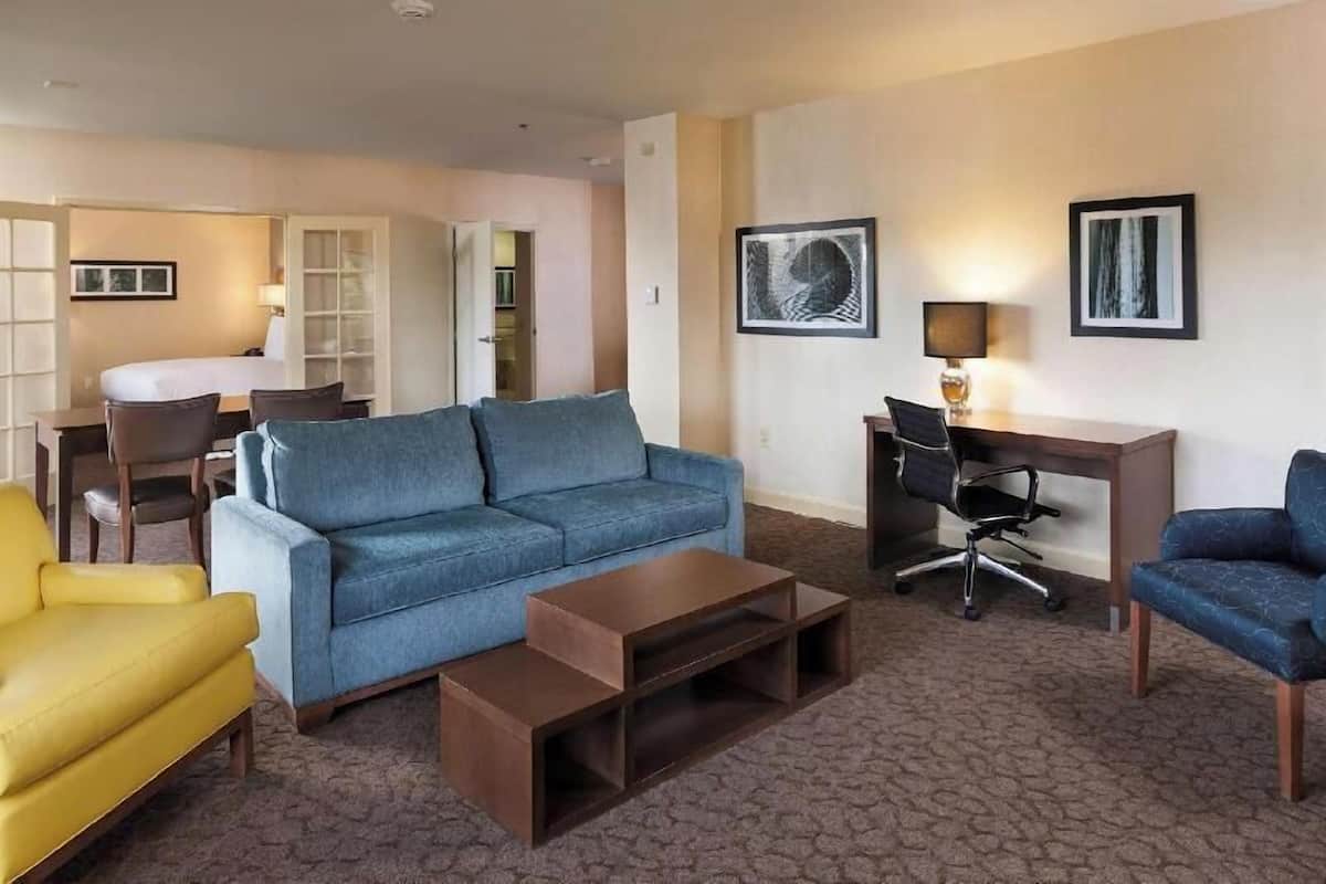 A hotel room with a blue sofa, a yellow armchair, a wooden coffee table, a desk with a chair, and two framed pictures on the wall.