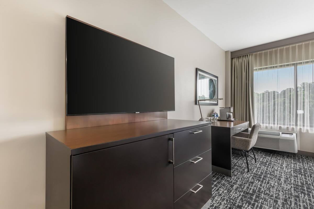 A hotel room with a large flat-screen TV mounted on a wooden cabinet, a desk with a chair, a coffee maker, and a large window with curtains.