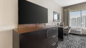 Une chambre d’hôtel dotée d’un grand téléviseur à écran plat fixé à un meuble en bois, d’un bureau avec une chaise, d’une machine à café et d’une grande fenêtre avec des rideaux.