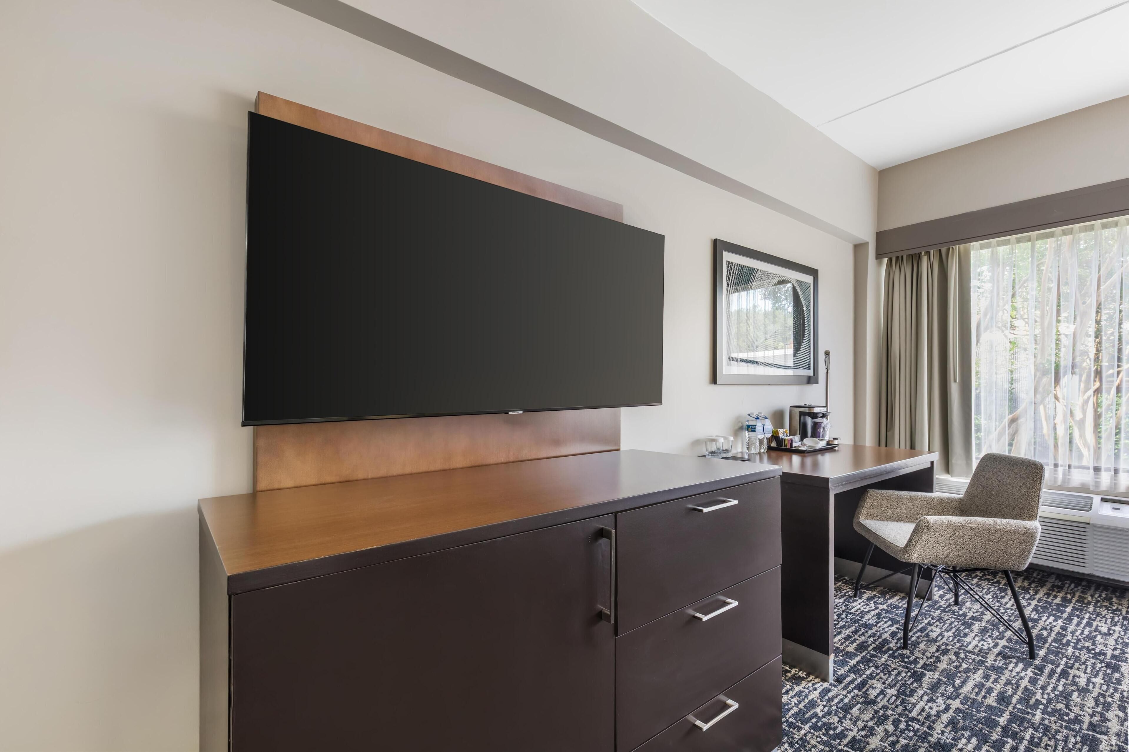 Hotelkamer met een flatscreen televisie bevestigd aan een houten kast, een bureau met stoel en een groot raam met gordijnen.