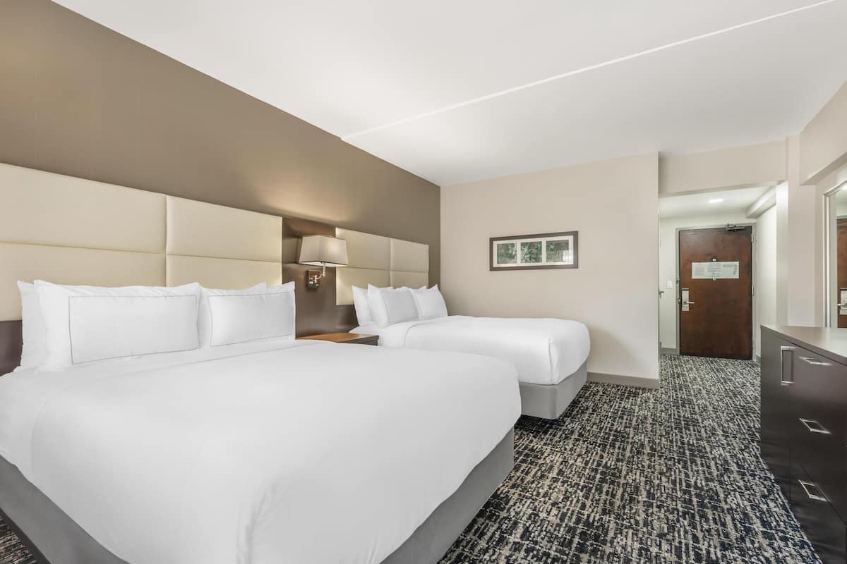 A hotel room with two beds, a bedside table with a lamp, a framed picture on the wall, and a door leading to another room.