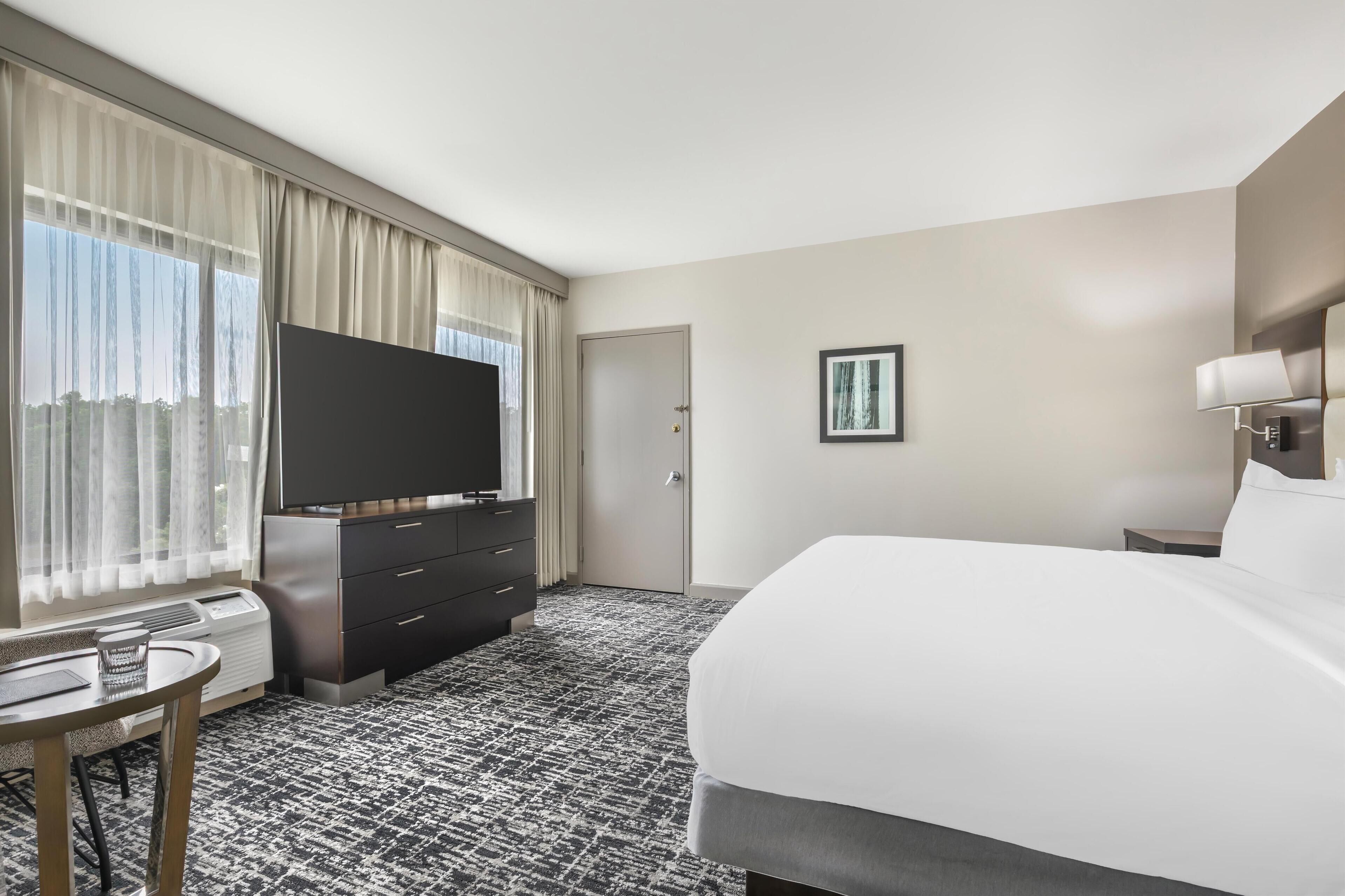 Habitación de hotel con cama, televisor de pantalla plana, escritorio con silla, cómoda y ventana con cortinas.