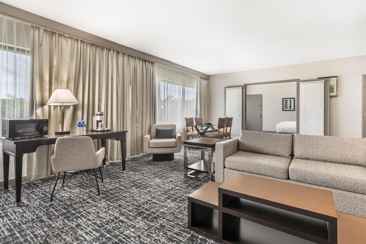 A hotel room with a large window, a sofa, a desk with a lamp, a TV, and a coffee machine.