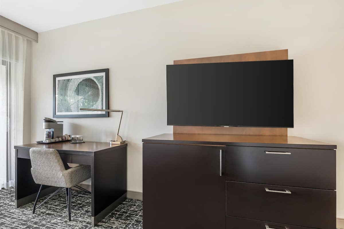 A hotel room with a dark wooden desk, a modern chair, a flat-screen TV, a framed artwork, and a coffee maker.