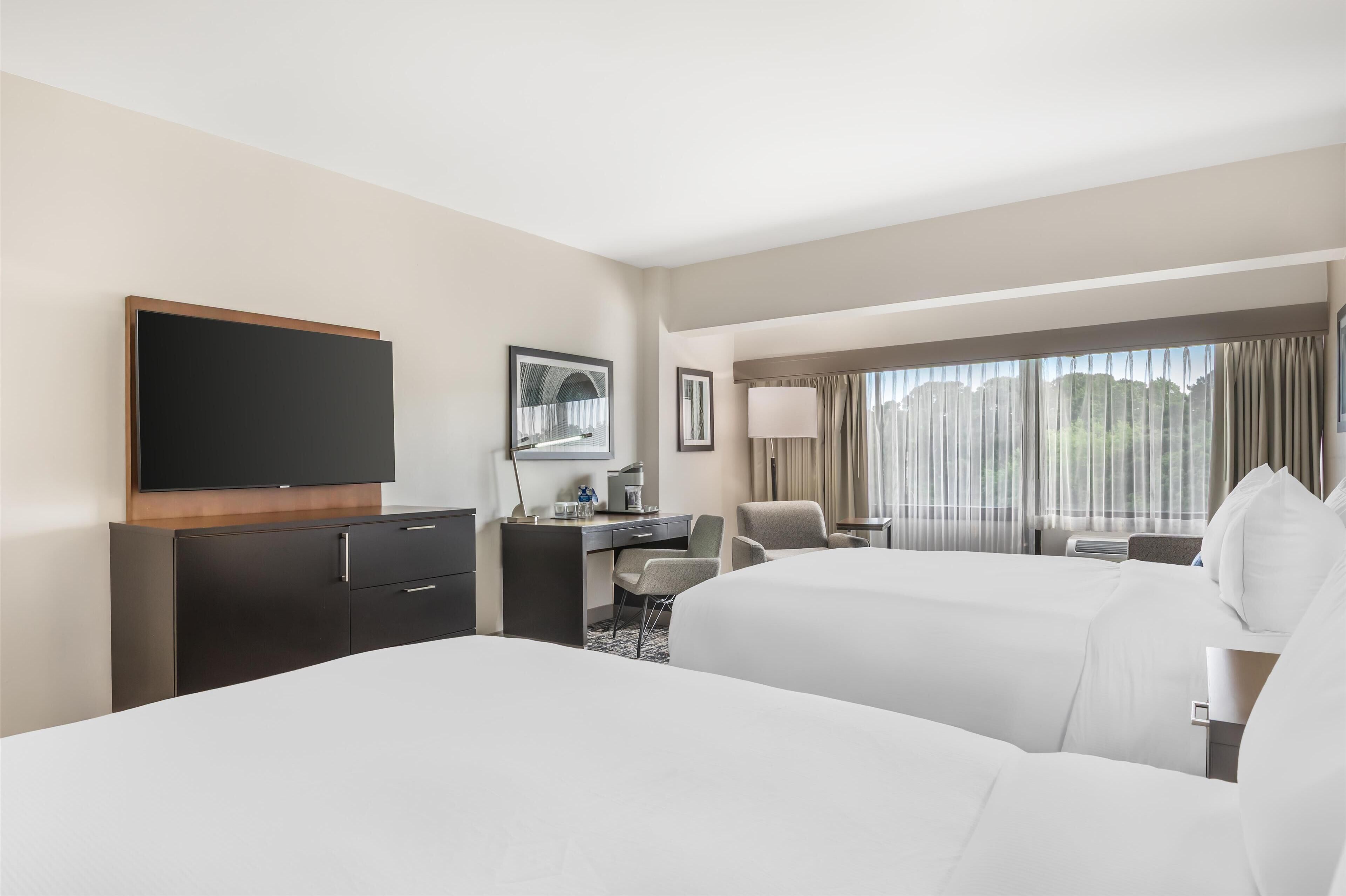 A hotel room with two beds, a desk, a television, and a large window with curtains.