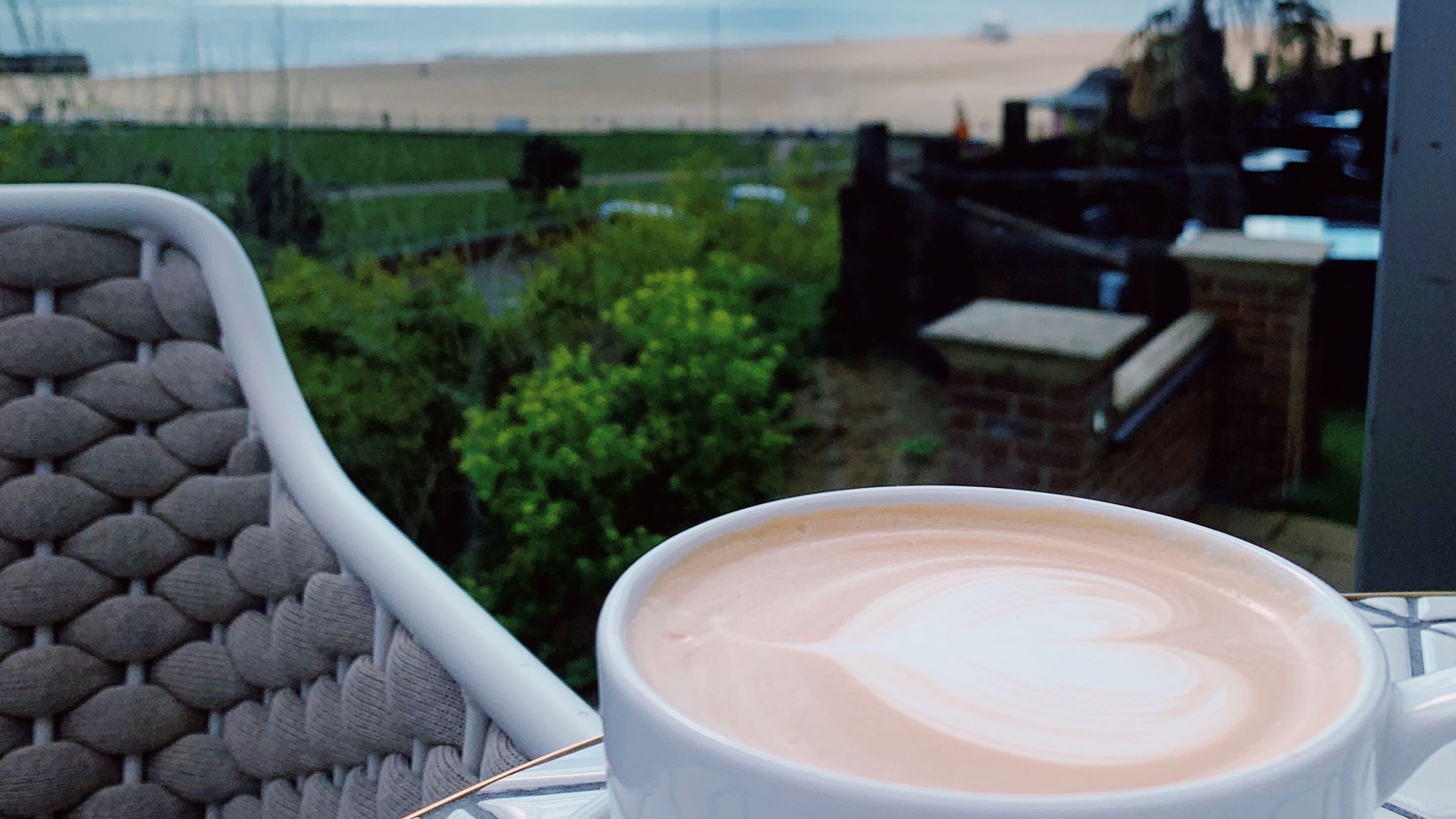 Eine Tasse Kaffee mit herzförmigem Latte-Art auf einem Tisch, mit Blick auf einen Garten und eine Strandansicht sowie einen Zaun.
