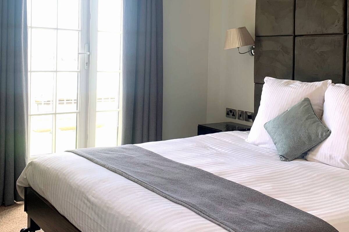 A hotel room with a large bed, a grey throw blanket, white pillows, and a grey accent pillow. There is a window with curtains, a bedside table with a lamp, and a wall-mounted light fixture.