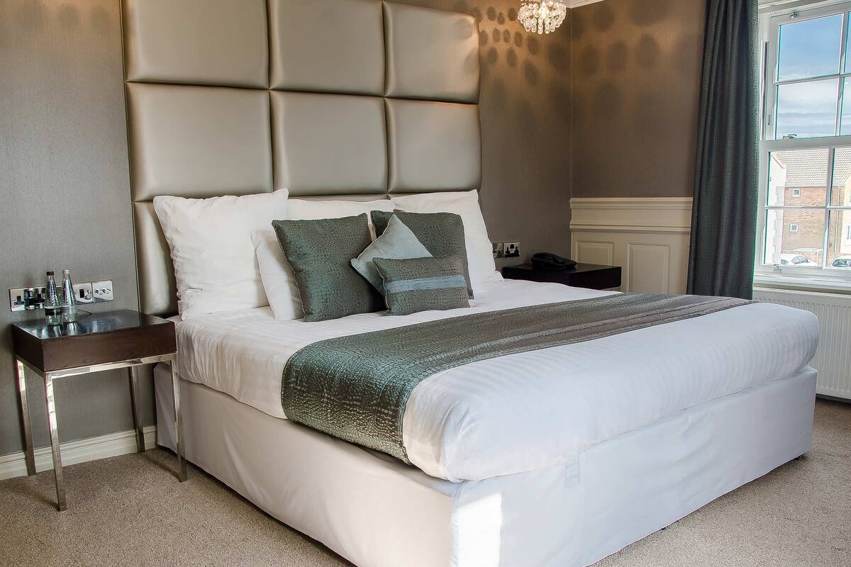 A neatly made bed with a headboard, a bedside table with a lamp, and a window with curtains.